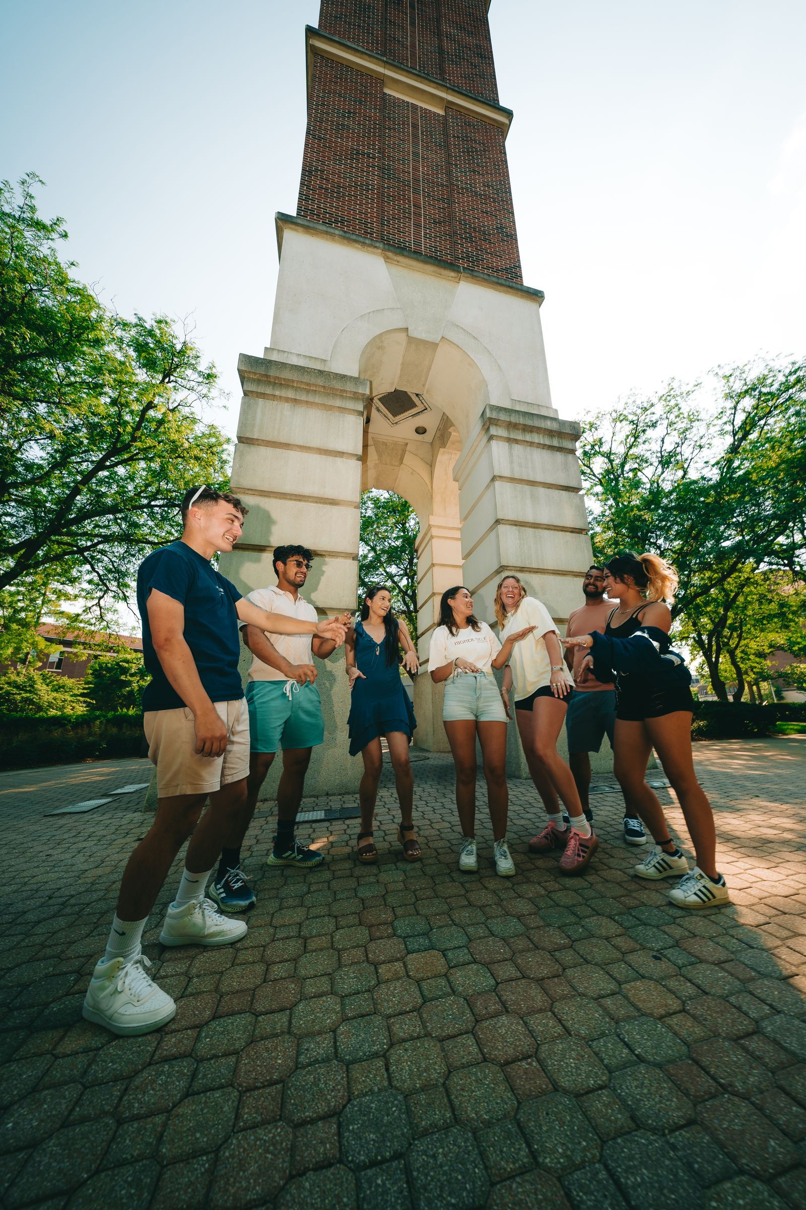 A group of seven people stands in a circle, chatting and laughing under a tall brick and stone tower.