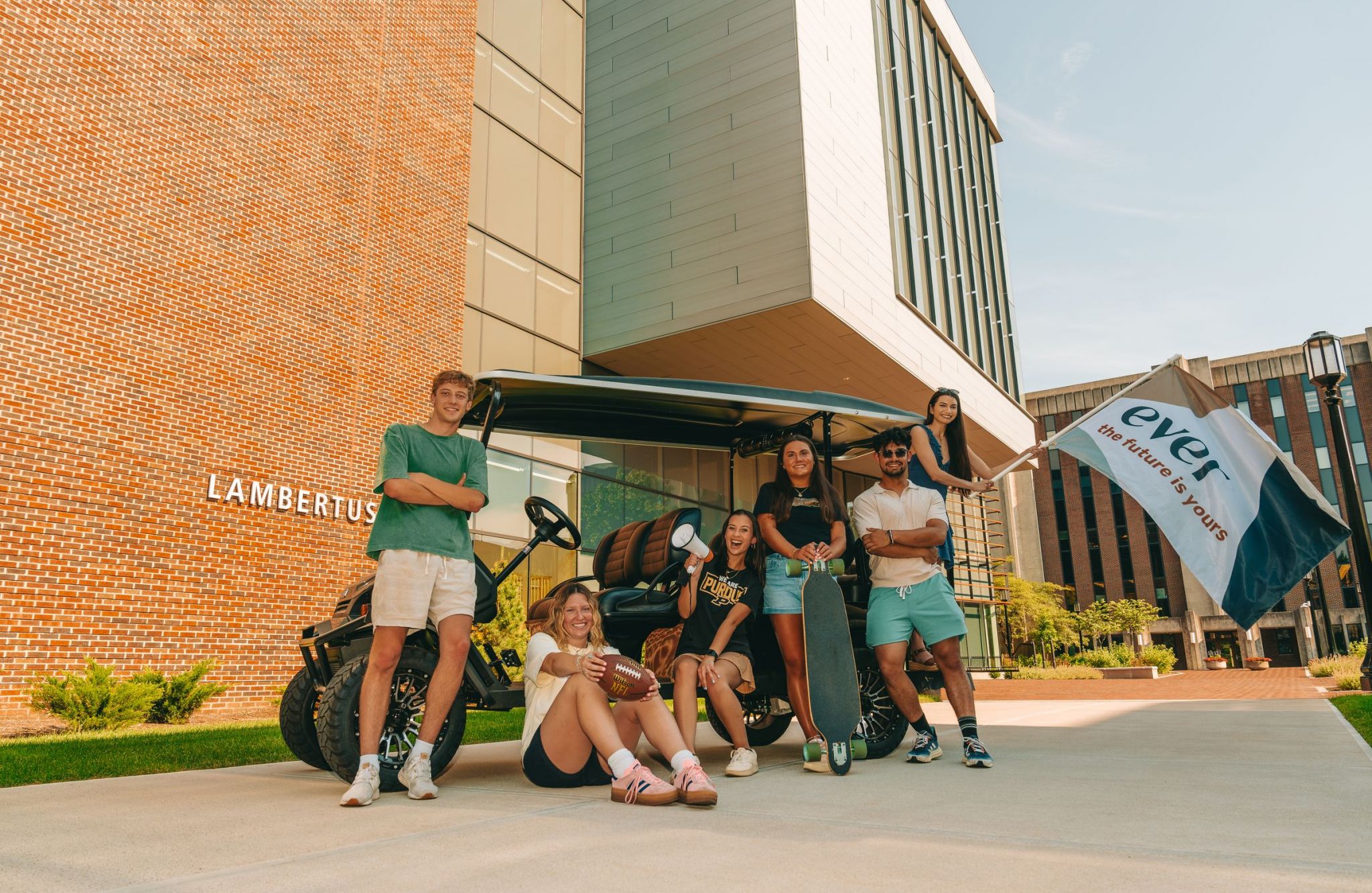 Group of six students pose with a golf cart and a flag outside a modern brick building labeled "LAMBERT.