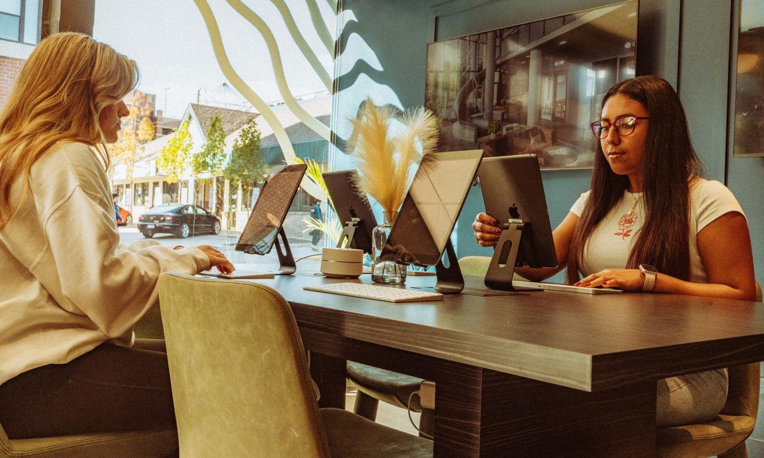 Two women sitting at a table in a modern workspace, working on laptops, with a street view seen through a window.