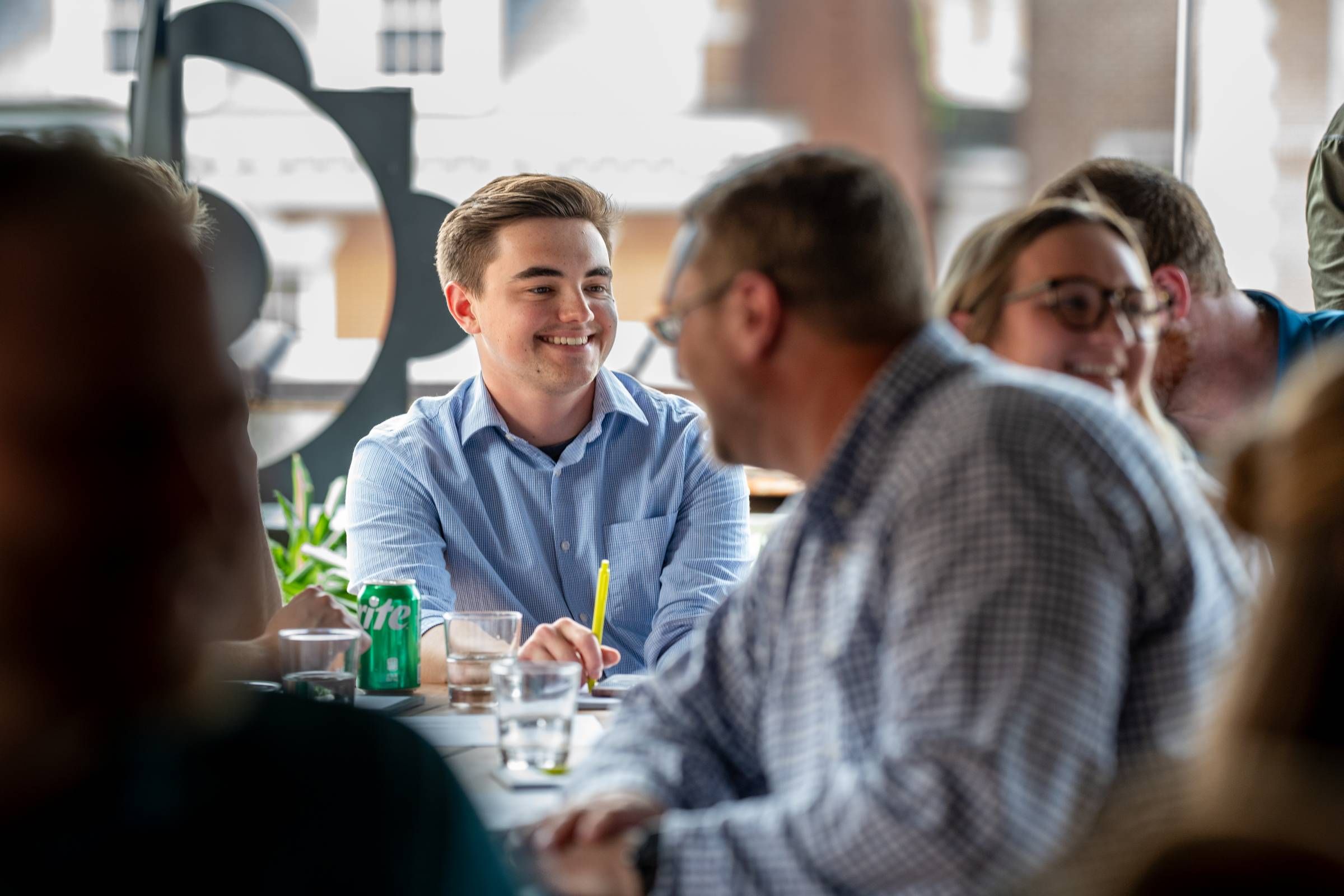 A group of people sitting at a table in a lively conversation, with drinks, including a Sprite can, on the table.