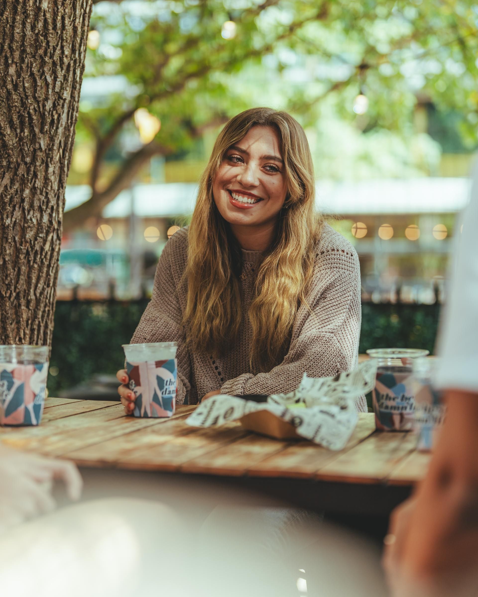 A woman with long hair is sitting at an outdoor table, smiling, holding a drink, with trees in the background.