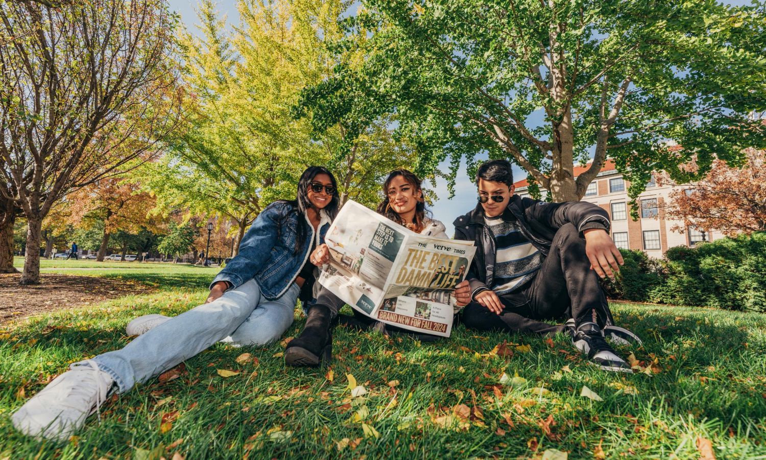 Three friends sit on grass reading a newspaper in a park with autumn trees in bright sunlight.