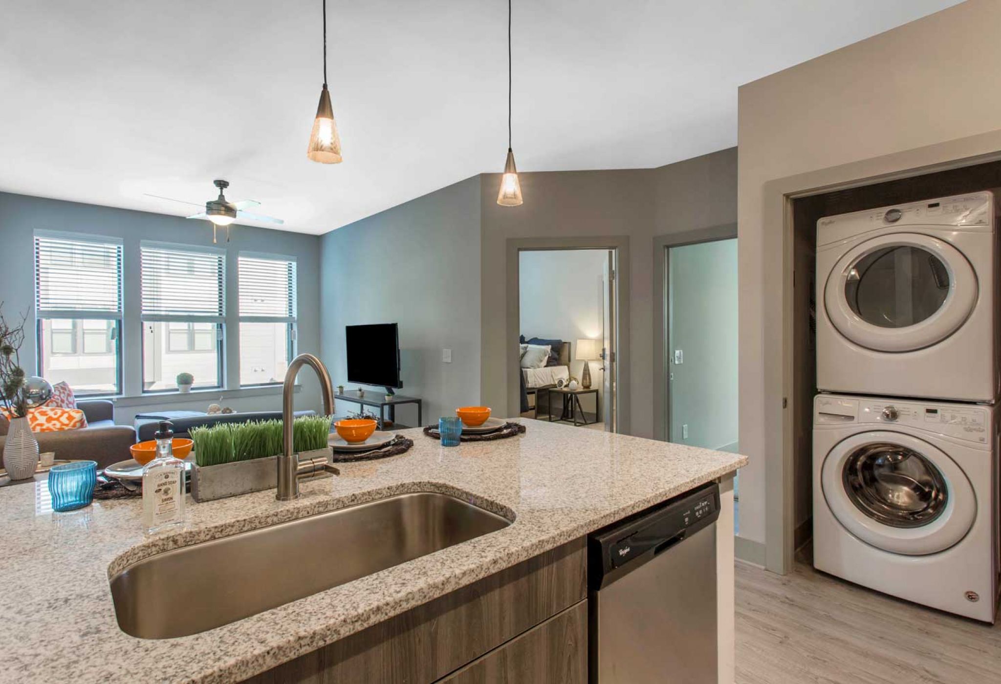 Discover the sleek 114 Earle modern apartment kitchen, featuring a granite island, pendant lights, a stacked washer and dryer, and a cozy living area in the background.