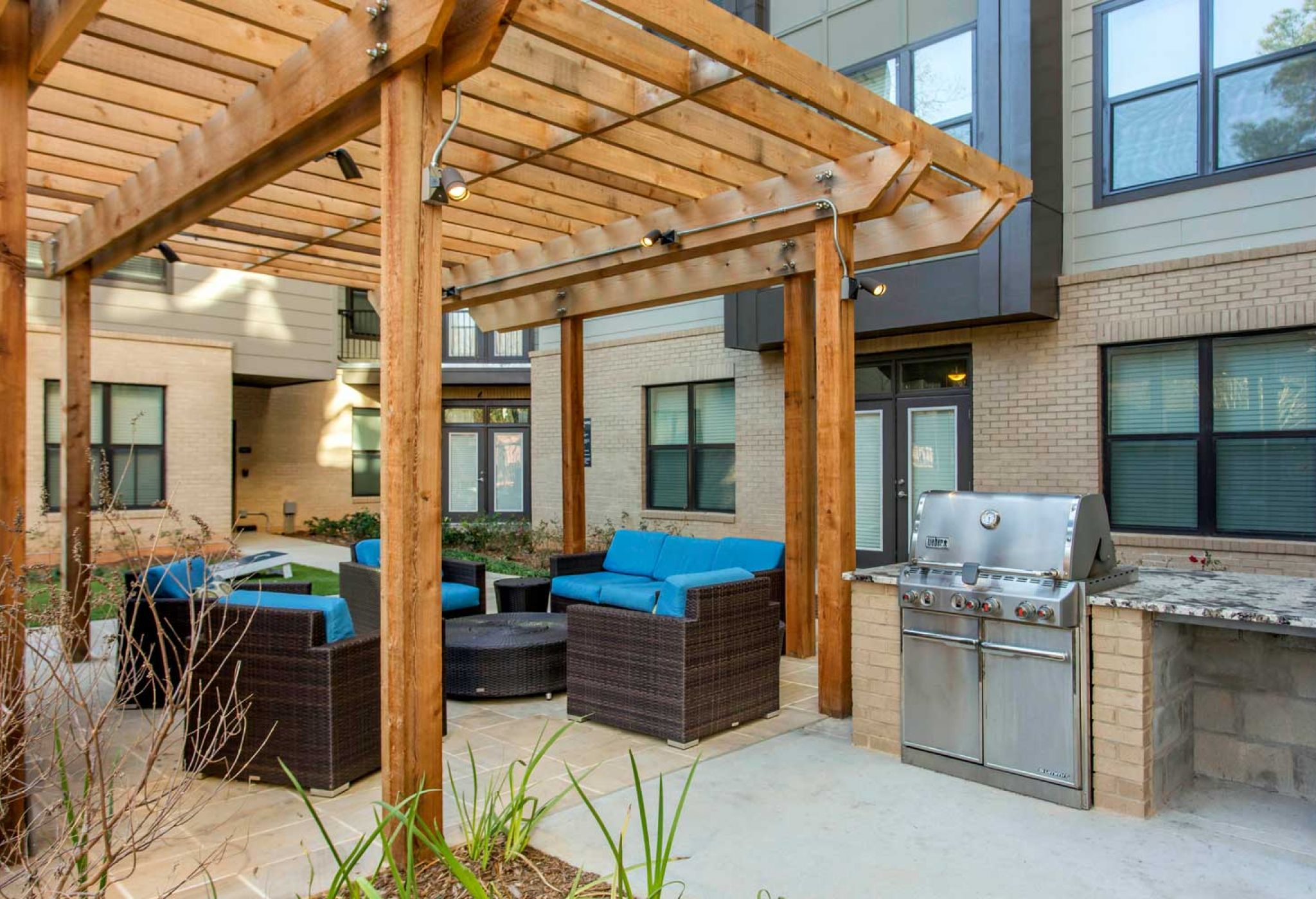 The outdoor patio at 114 Earle features a wooden pergola, blue cushioned seating, and a stainless steel grill beside a brick building.