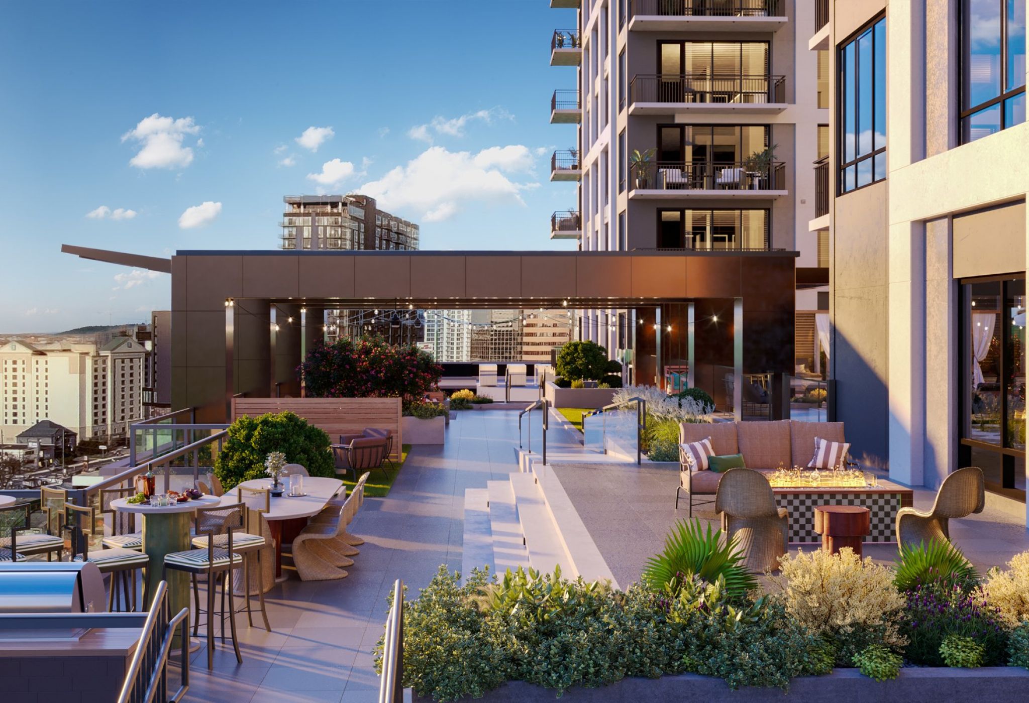 A modern apartment building rooftop terrace in Nashville featuring seating areas, tables, and lush greenery. This LOCAL gem offers panoramic city views under a clear blue sky, with additional high-rise buildings visible in the background.