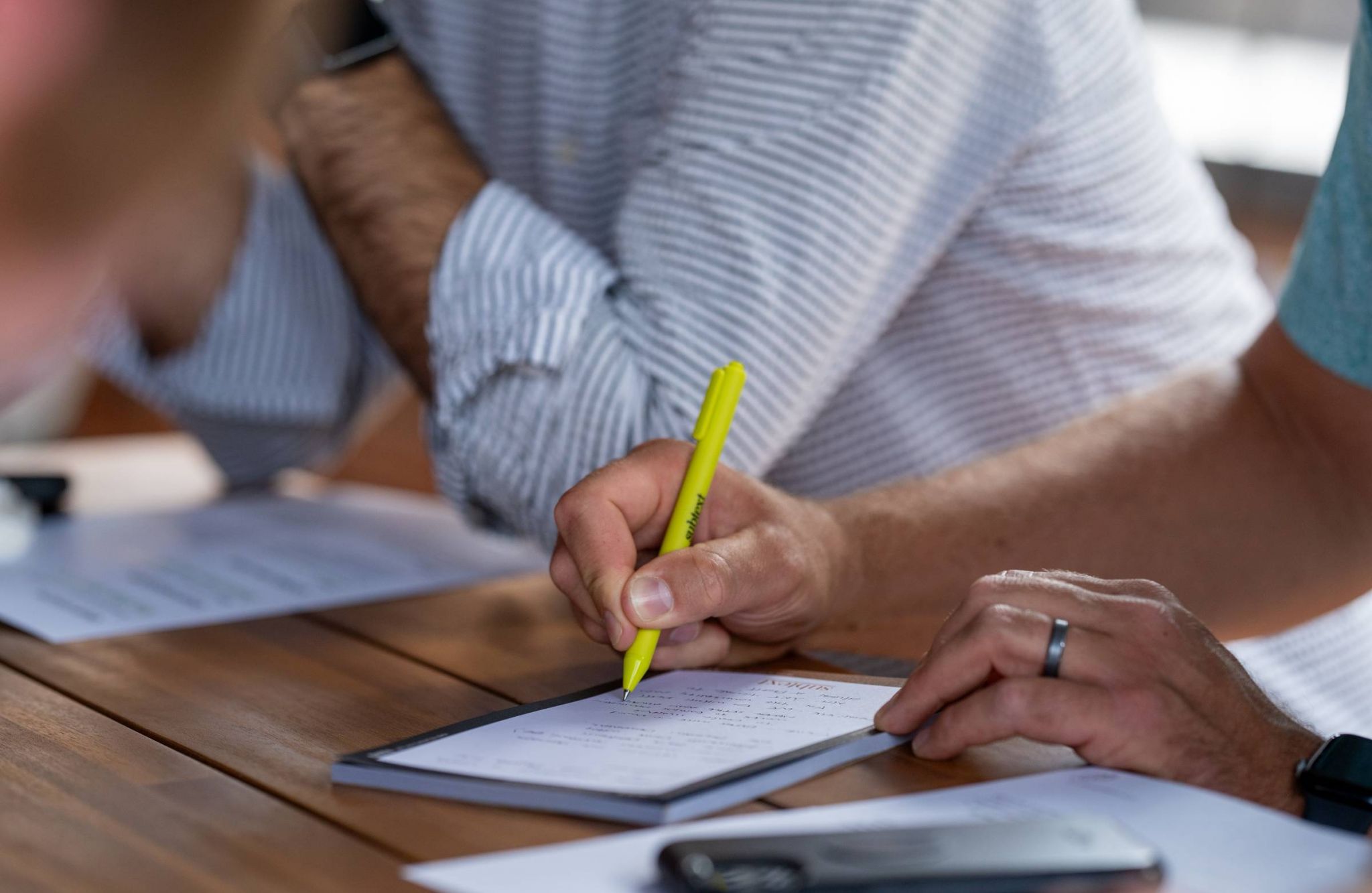 Close-up of two people writing notes with a yellow highlighter on a notepad, sitting at a wooden table.