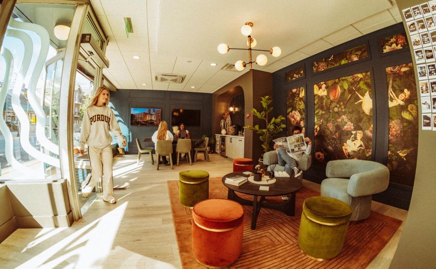 Cozy interior with vibrant decor, colorful seating, and people lounging and chatting. A woman enters through the doorway.