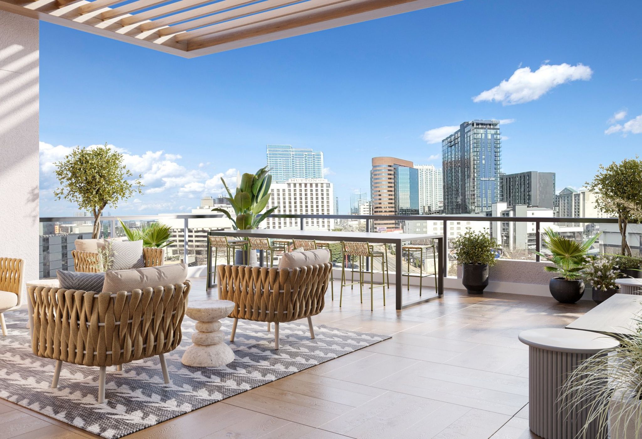 A modern rooftop terrace in Nashville features cozy wicker chairs arranged around a small stone table on a patterned rug. Potted plants are scattered throughout, with a stunning cityscape and blue sky visible in the background. A wooden pergola provides partial shade to this local gem.