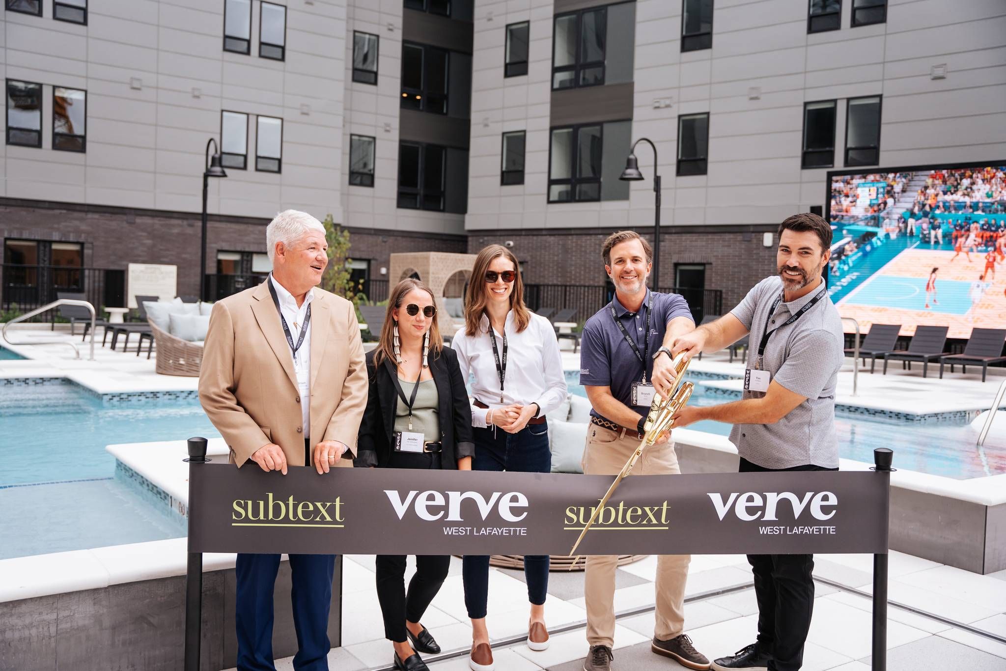 Five people stand by a ribbon cutting ceremony at a pool area with a "Verve West Lafayette" banner and large building behind, hinting at premium student housing near Purdue University.
