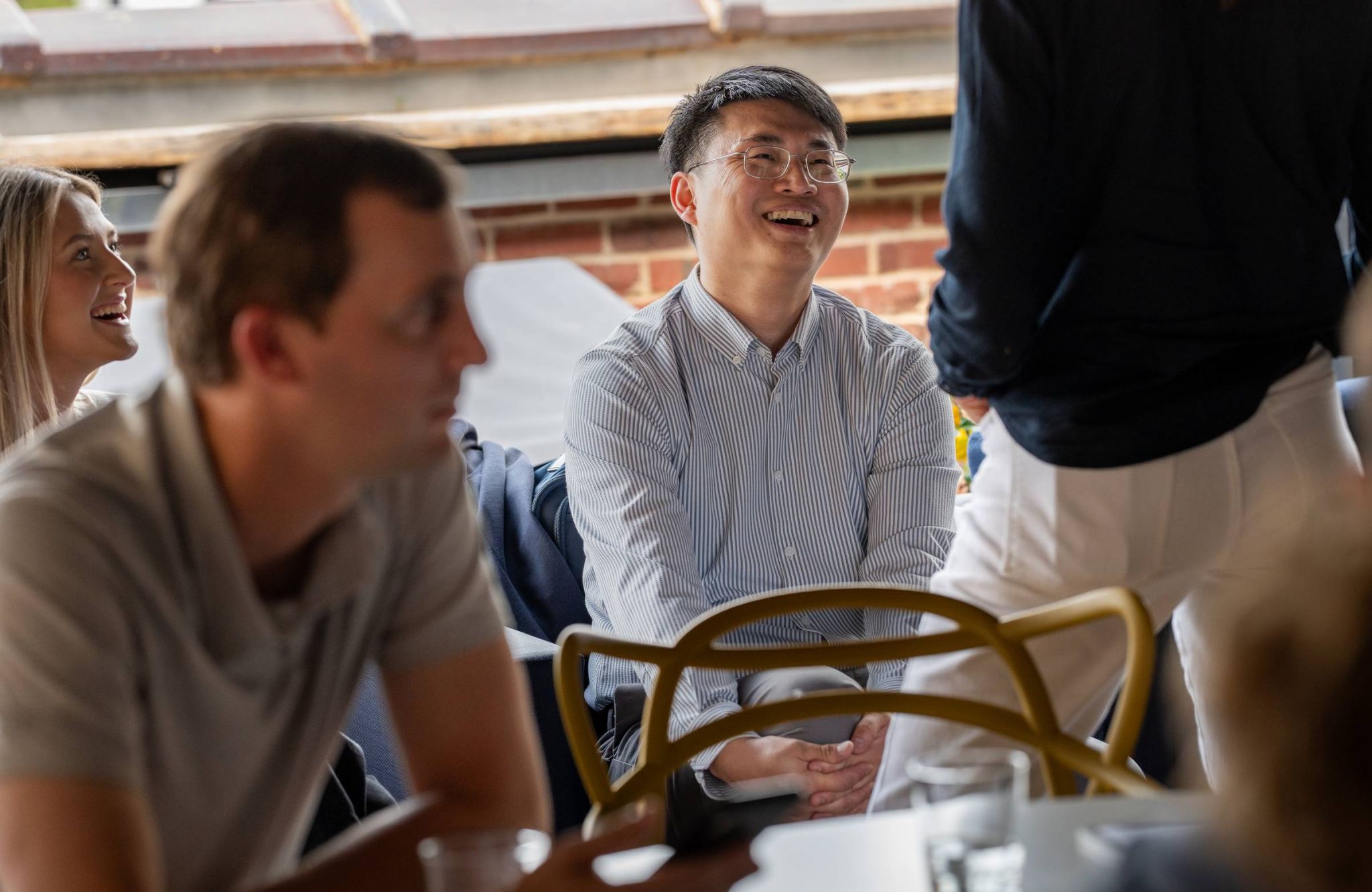 A man wearing glasses and smiling while talking to others in a casual indoor setting.