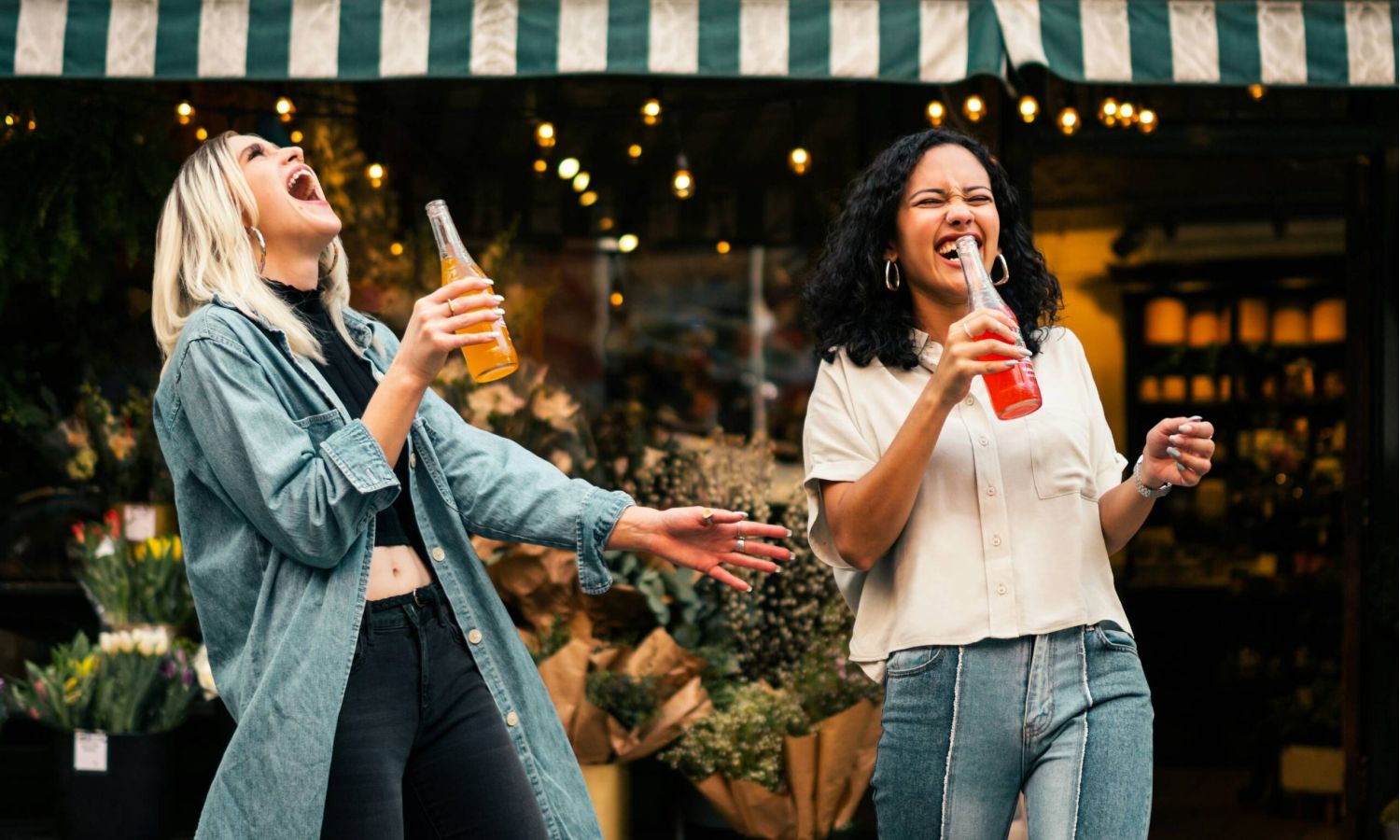 Two women laughing and sipping soda outside Subtext Living, with string lights casting a glow over vibrant bouquets.