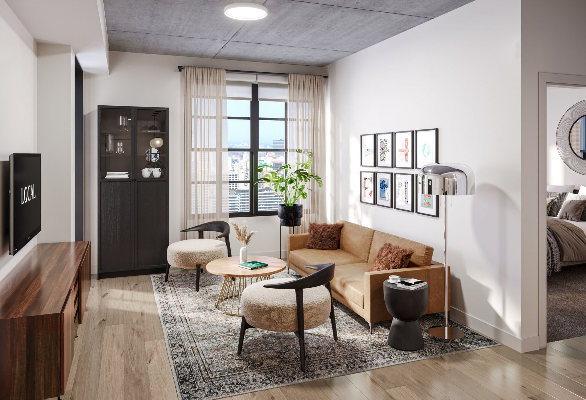 Modern living room with a minimalist design. Features a tan sofa with brown cushions, a round wooden coffee table, two beige cushioned chairs, and a tall plant. A floor lamp stands near the sofa. The room has light wood flooring and a large window with sheer curtains. Adjoining bedroom visible.