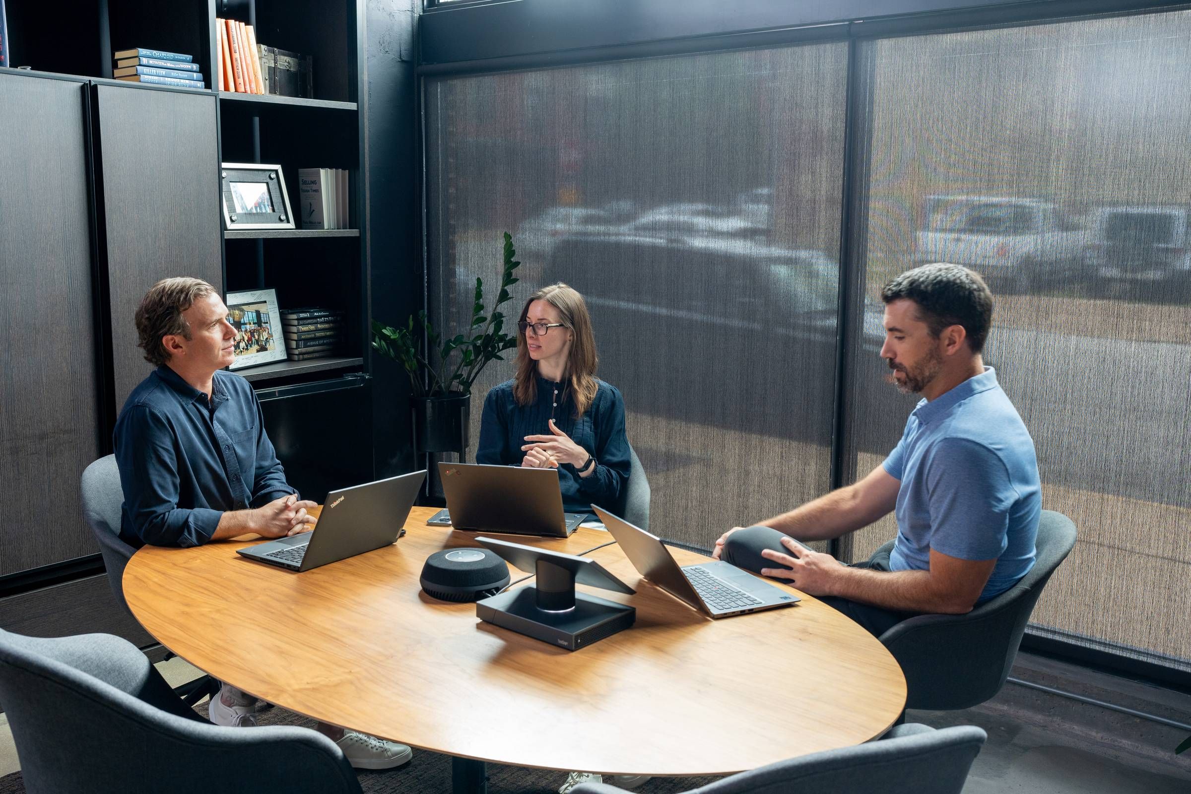 Three people having a meeting around a table with laptops and notebooks in a modern office setting with large windows.