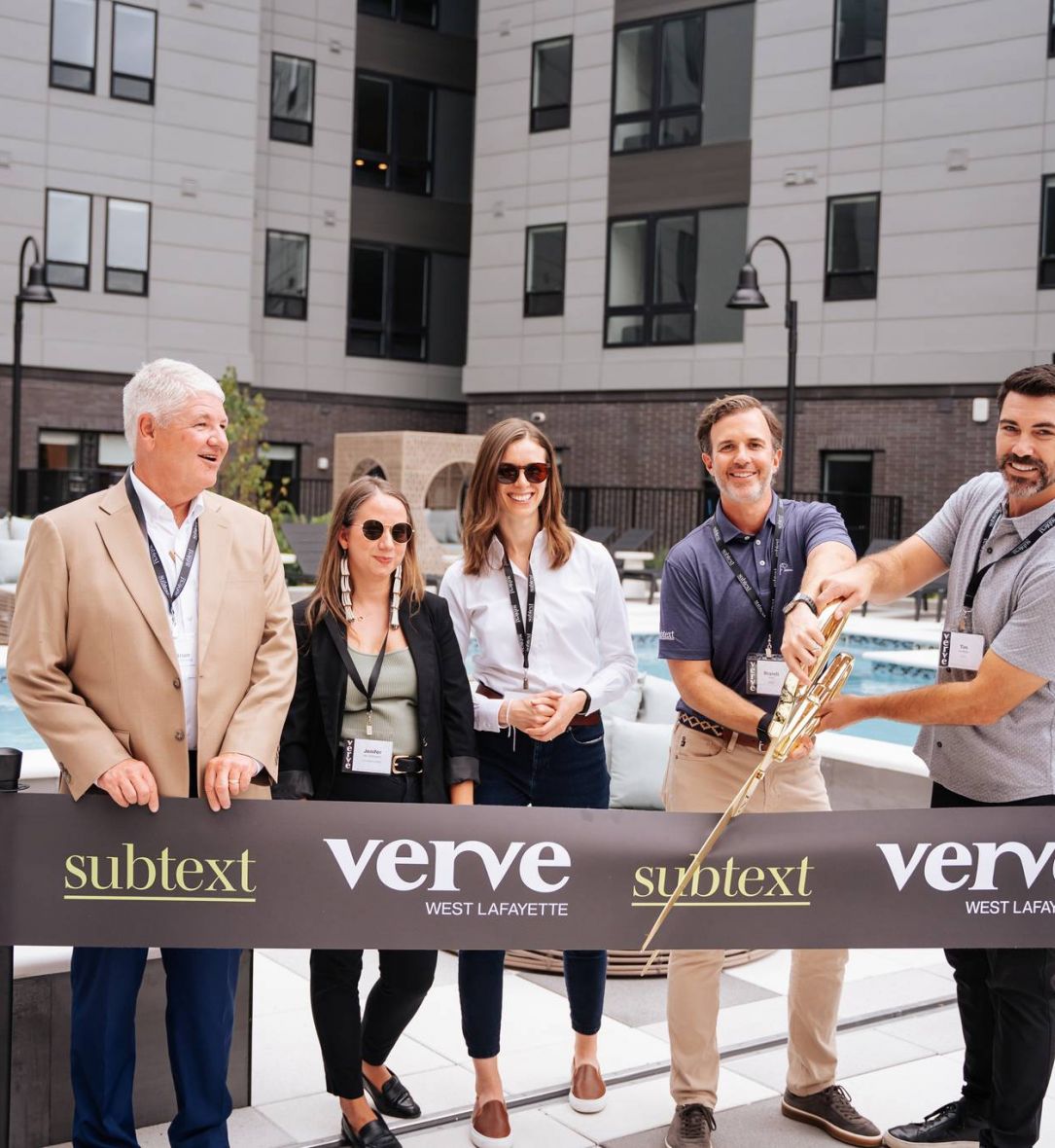 Five people stand by a ribbon cutting ceremony at a pool area with a "Verve West Lafayette" banner and large building behind, hinting at premium student housing near Purdue University.