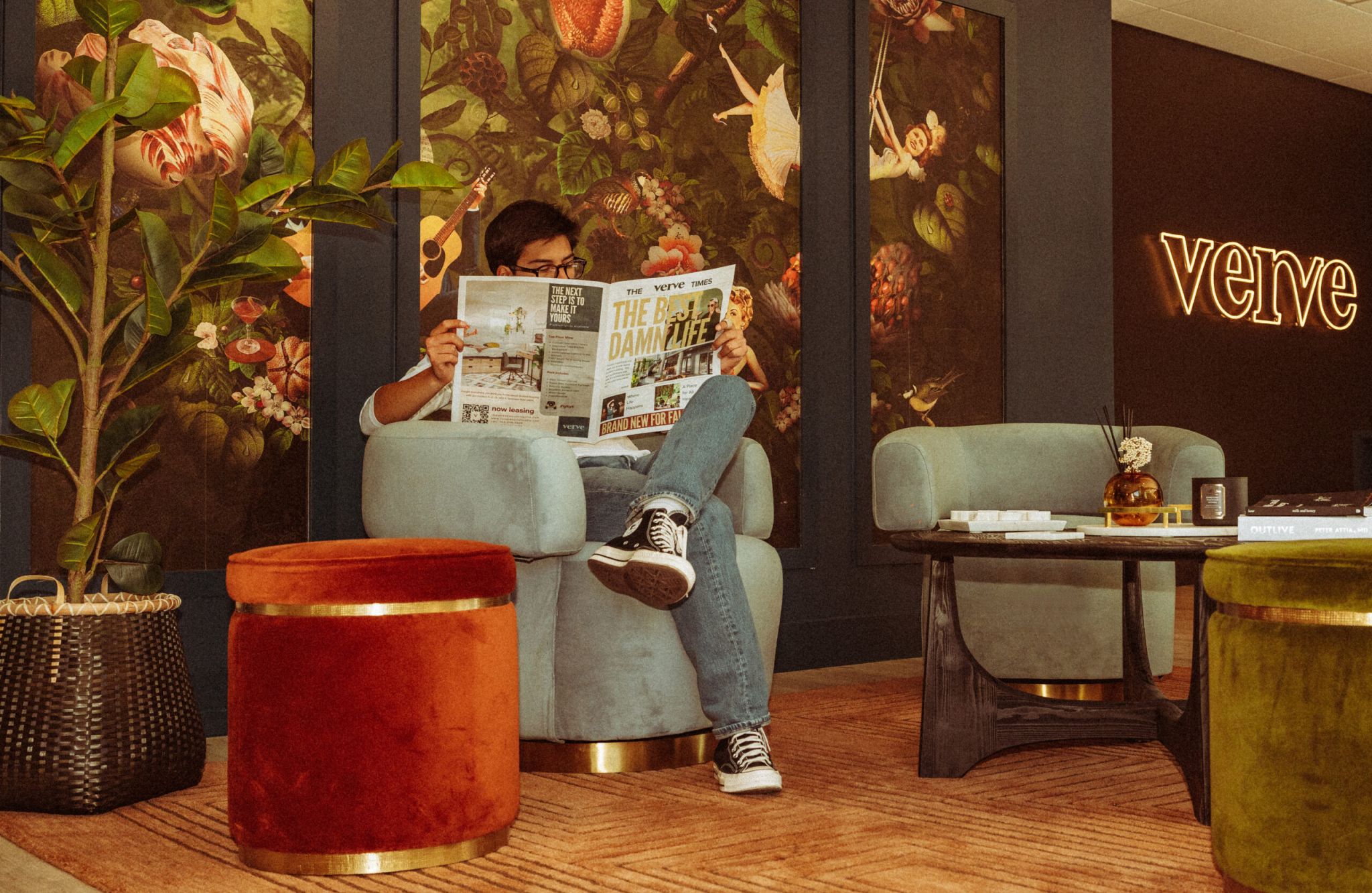 Person sitting in a cozy, stylish room reading a newspaper with vibrant floral wallpaper and neon "verve" sign in the background.