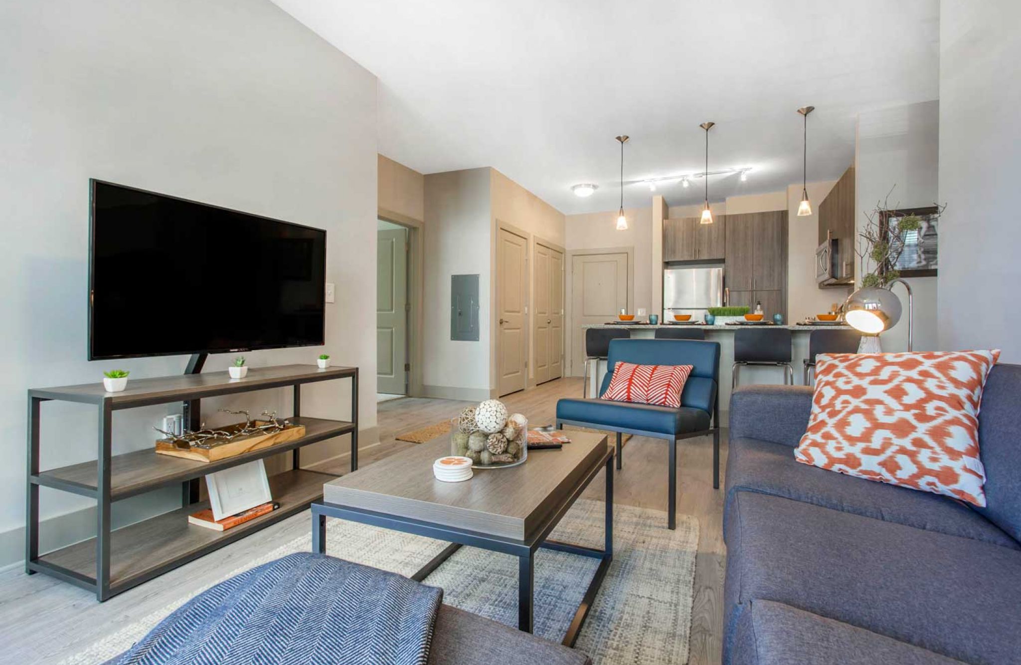 Modern living room at 114 Earle features a blue sofa, patterned pillows, large TV on stand, and an open kitchen in the background with pendant lights.