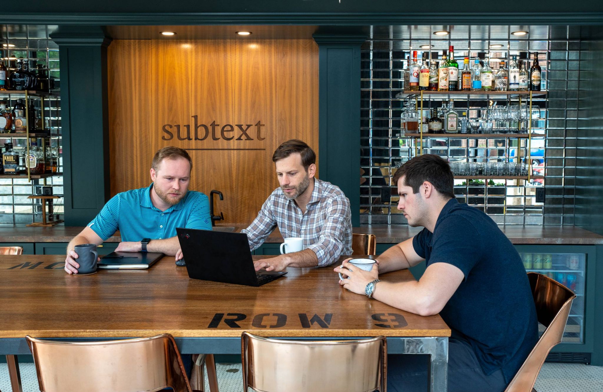 Three men sitting at a table working on a laptop in a bar setting with a "subtext" sign in the background.