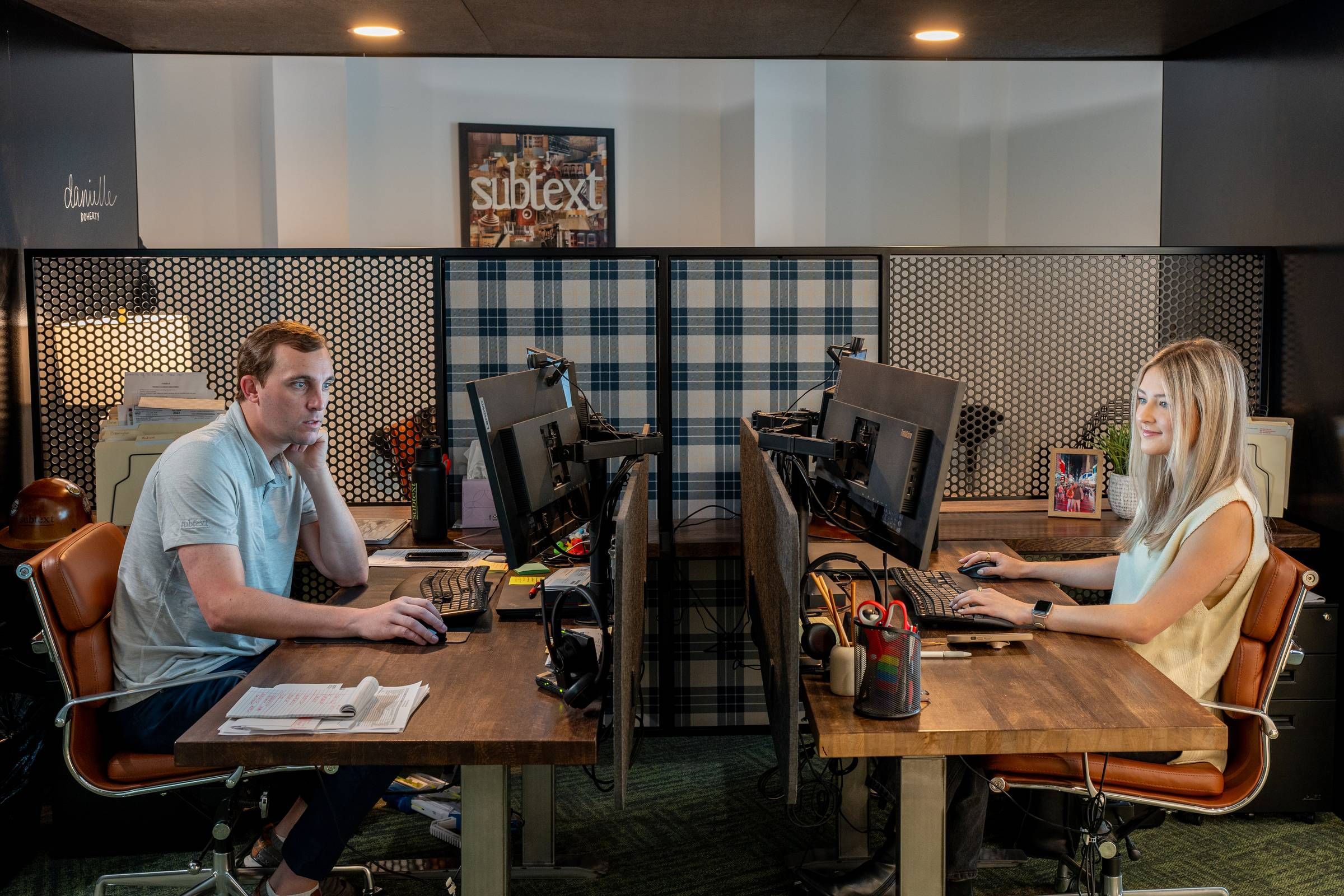 Two people working at adjacent desks with computers in a well-lit office, separated by a checkered partition.