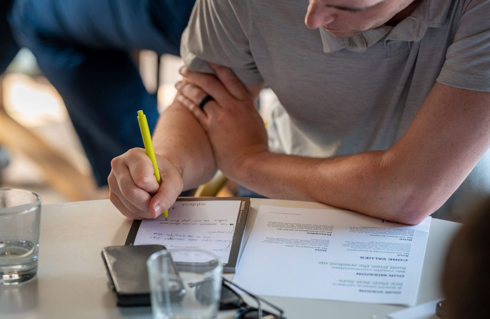 Close-up of two people writing notes with a yellow highlighter on a notepad, sitting at a wooden table.