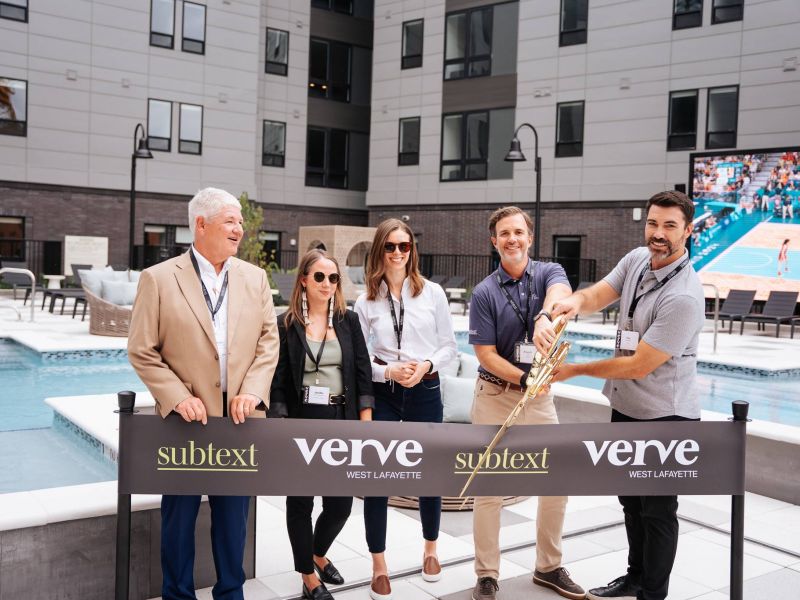 Five people stand by a ribbon cutting ceremony at a pool area with a "Verve West Lafayette" banner and large building behind, hinting at premium student housing near Purdue University.