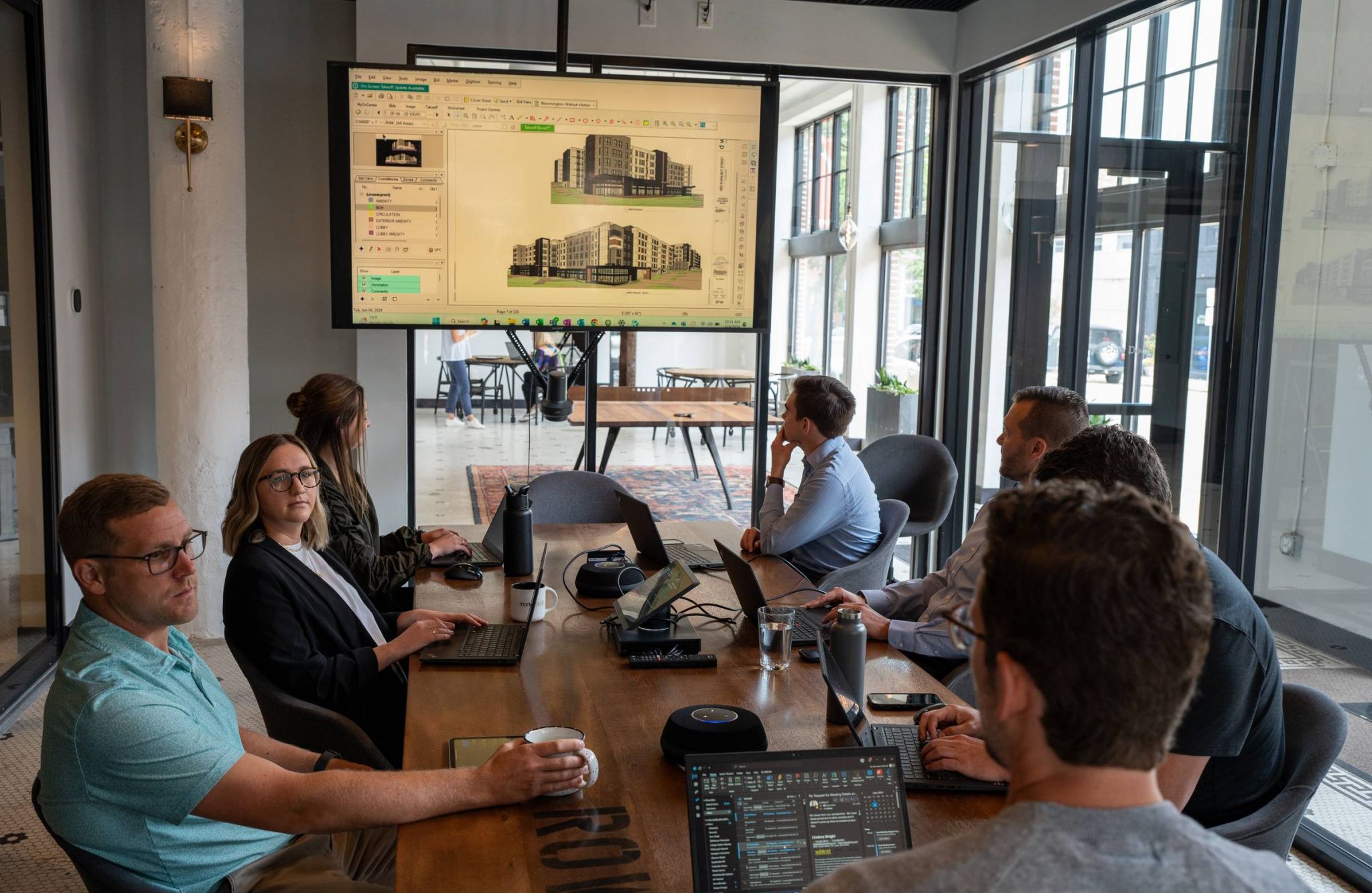 A group of people in a modern office having a meeting, with architectural designs displayed on a screen.