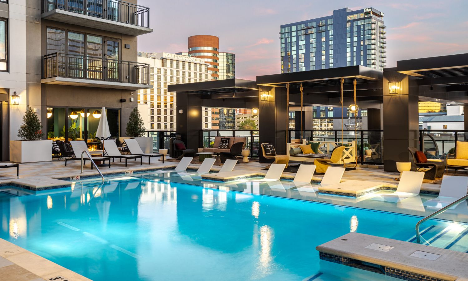 Subtext Living: Enjoy a rooftop patio with wicker chairs, lush plants, and a wooden pergola, all overlooking the city skyline on a sunny day.