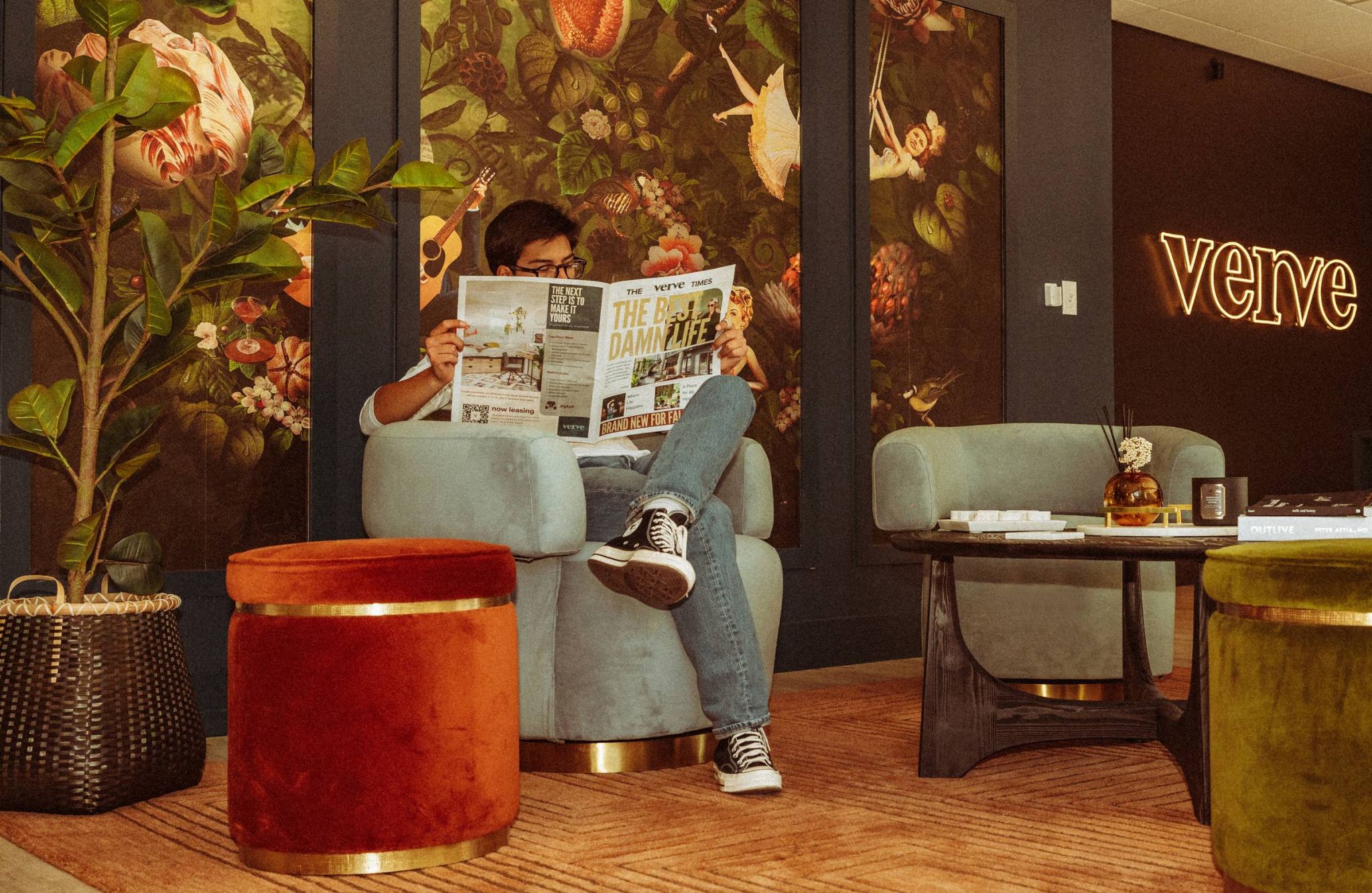 Person sitting in a cozy, stylish room reading a newspaper with vibrant floral wallpaper and neon "verve" sign in the background.