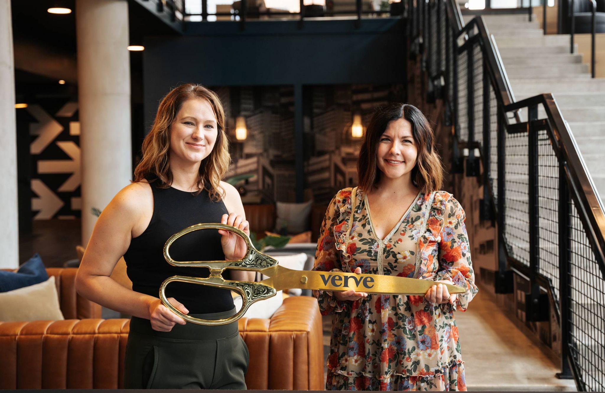 Two women smile, holding a giant pair of ceremonial scissors labeled "Verve" in a modern indoor setting.