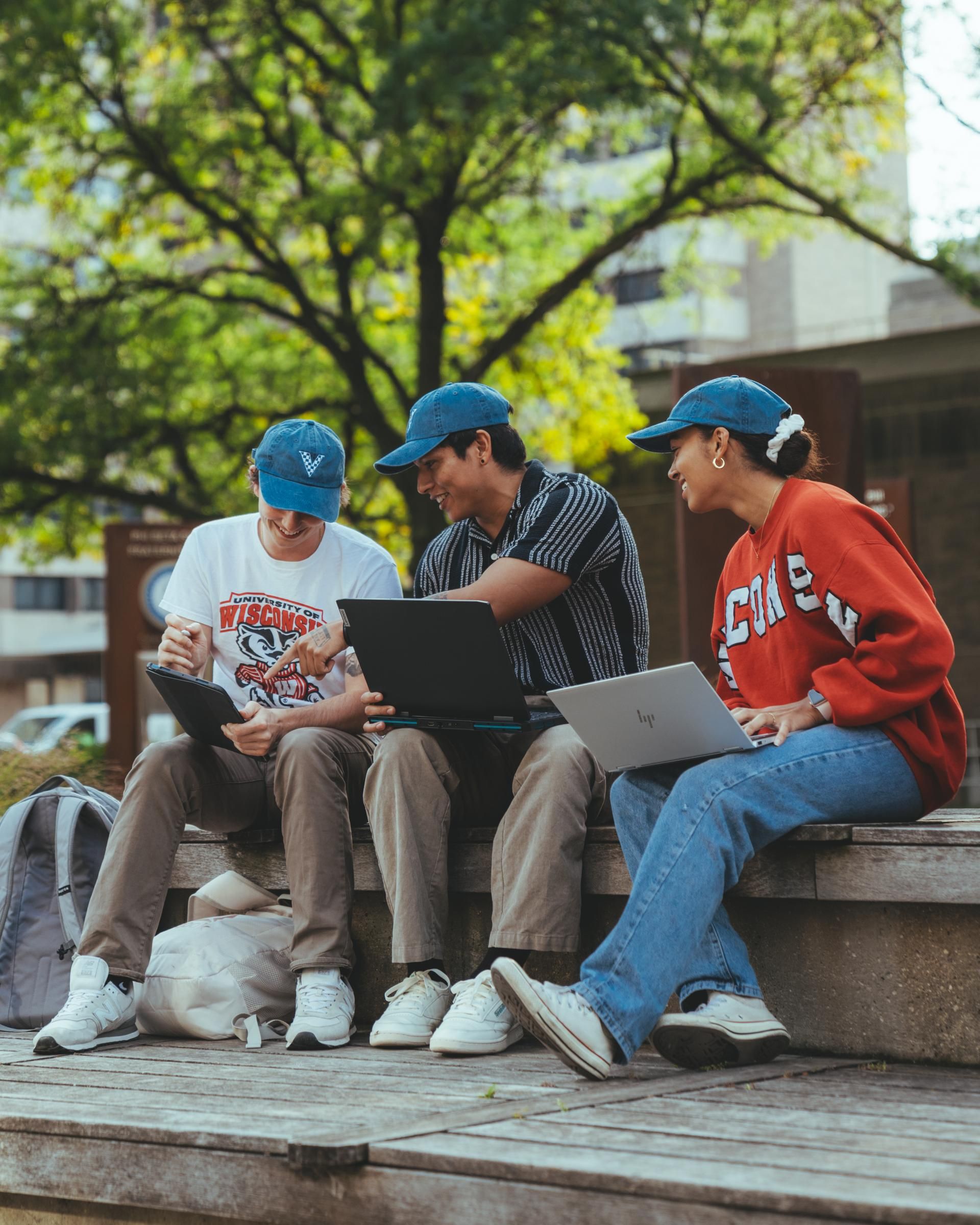 Three people in casual attire sit on a wooden bench outdoors, interacting with laptops and smiling.