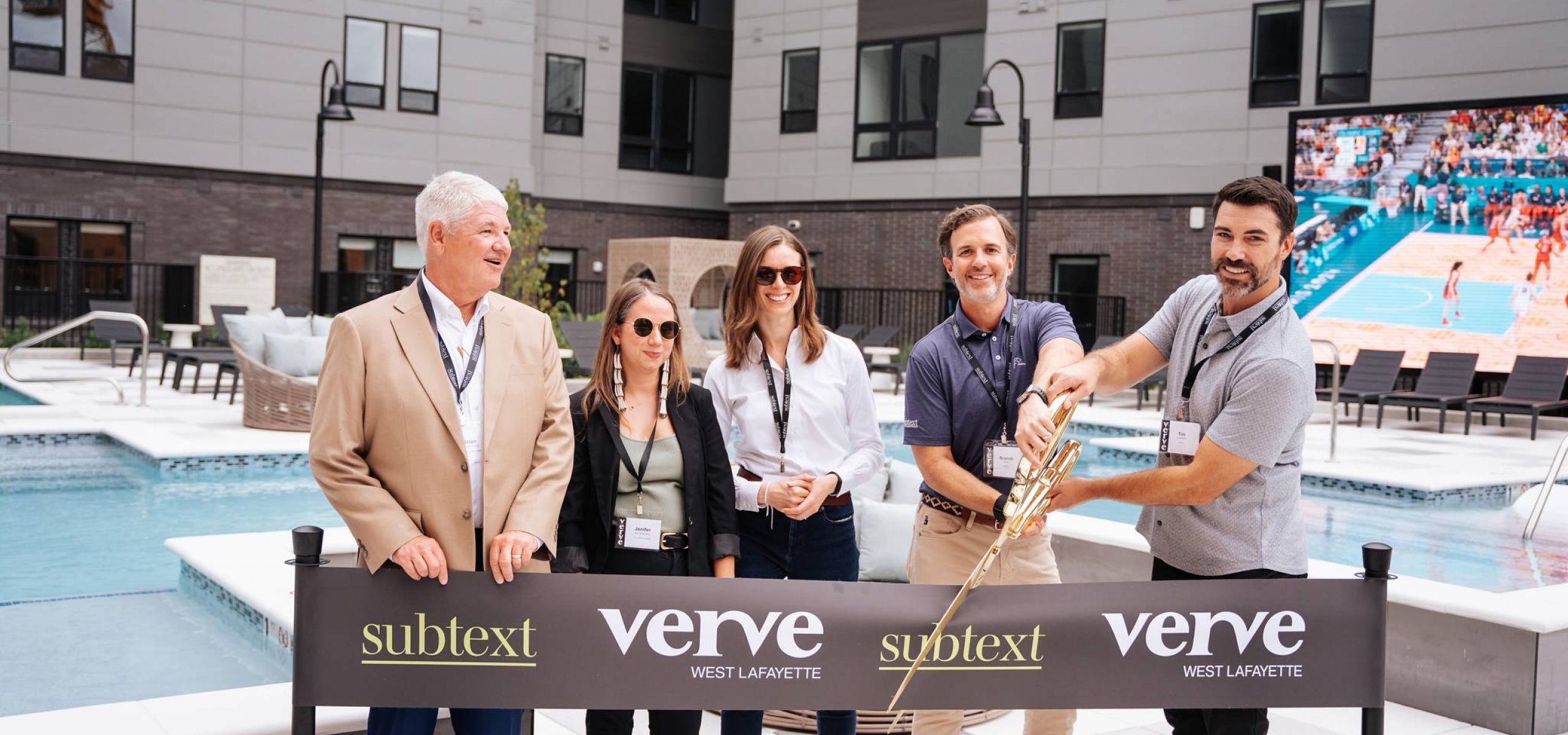 Five people stand by a ribbon cutting ceremony at a pool area with a "Verve West Lafayette" banner and large building behind, hinting at premium student housing near Purdue University.