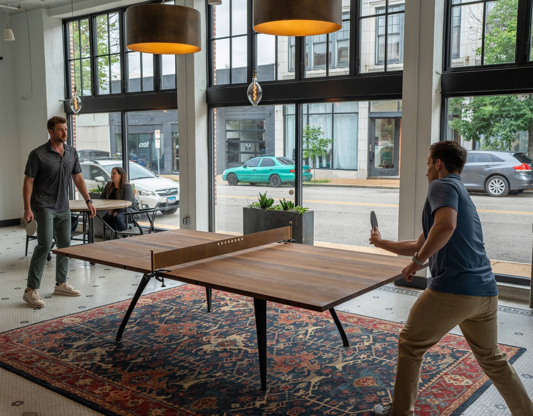 Two men playing ping pong in a modern room with large windows, while a woman watches from a table on the side.